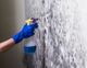 Hand in blue glove spraying moldy wall with a cleaner.