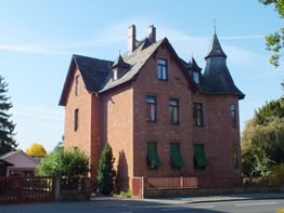 Backsteinvilla mit Türmchen und grünen Fensterläden, umgeben von Bäumen an einer ruhigen Straße.