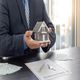 Man in suit holding house model over insurance papers and keys on a desk.