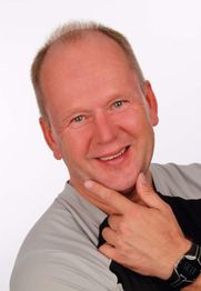 Ein lächelnder Mann mit kurzen, hellen Haaren posiert mit der Hand am Kinn vor weißem Hintergrund.