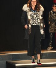 Frau auf Laufsteg mit gemustertem Outfit und Pelzmantel, im Hintergrund Person in Leopardenmuster.