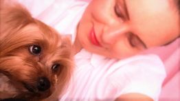 Eine Frau lächelt und kuschelt mit einem kleinen Hund in einem rosa beleuchteten Raum.