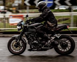 Motociclista de preto pilotando moto esportiva em movimento, fundo urbano desfocado.