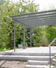Terrassenüberdachung mit Glasschiebedach im Garten, umgeben von Bäumen und Pflanzen.