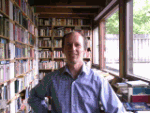 Ein Mann steht in einem Raum voller Bücherregale, neben einem großen Fenster.