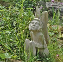 Steinfigur eines Mädchens mit Flügeln im Gras, umgeben von Pflanzen.