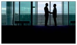 Zwei Personen geben sich im Schattenriss die Hand vor einem Fenster mit Blick auf einen Flughafen.
