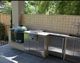 Outdoor kitchen with a grill, cabinets, and fridge, set against a tiled wall with greenery behind.