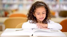 Young girl reading a book at a table in a library setting.