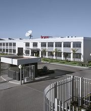 Edificio moderno con antena satelital y banderas; entrada cercada al frente.