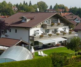 Großes Haus mit Balkon, grünem Garten und überdachtem Pool, umgeben von Bäumen und Gebäuden.