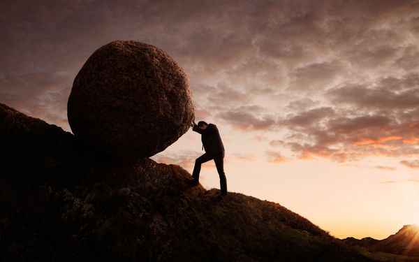 Ein Mann rollt symbolisch einen überdimensional großen Fels einen Hügel hinauf, um seine Willensstärke zu beweisen.