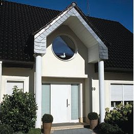 Eingang eines Hauses mit rundem Fenster, zwei Säulen und schwarzem Dach. Weiße Tür und Pflanzen.