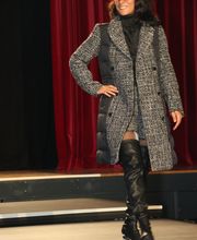 Frau auf Laufsteg mit grauem Mantel, roter Brille und schwarzen Stiefeln vor rotem Vorhang.