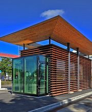 Modernes Gebäude mit Glaswänden und Holzlatten, umgeben von Parkflächen und blauem Himmel.
