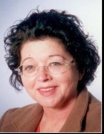 Frau mit Brille und lockigem, dunklem Haar lächelt in die Kamera. Sie trägt ein braunes Sakko.