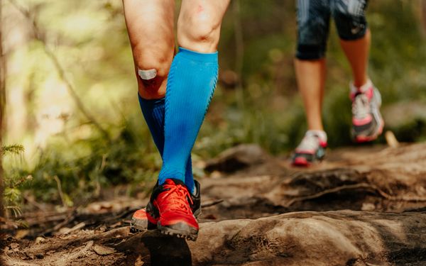 Sportlerin mit blauen Kompressionsstrümpfen läuft mit einer weiteren Person über einen unebenen Waldweg.