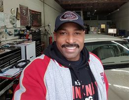 Man smiling in a garage, wearing a cap and sports jacket. Car and tools visible in background.