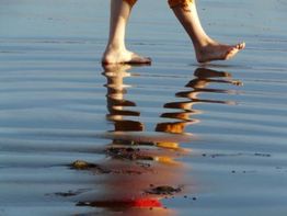 Füße eines Menschen laufen auf seichtem Wasser, reflektiert in den Wellen unter einem blauen Himmel.
