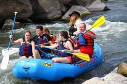 People in a blue raft navigating rapids, paddling enthusiastically, with a guide steering.
