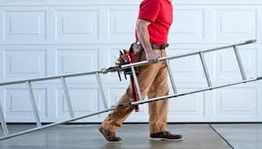 Person in red shirt and khakis carrying a ladder in front of a white garage door.