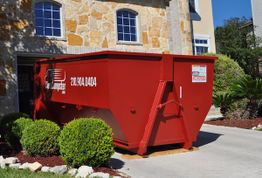 Red dumpster in a residential driveway beside a stone house with bushes in the foreground.