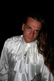 Mann mit langem Haar und weißem Rüschenhemd vor schwarzem Hintergrund.