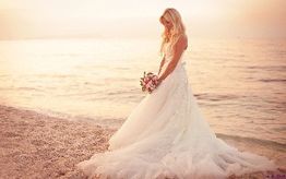 Braut in weißem Kleid am Strand bei Sonnenuntergang, hält einen Blumenstrauß, schaut aufs Meer.