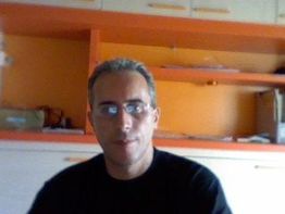 Uomo con occhiali in una stanza con mensole arancioni, seduto, guarda verso la fotocamera.