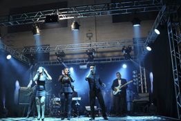 Banda se apresentando em palco iluminado com três cantores e guitarrista ao fundo.