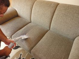 Pessoa limpando sofá cinza com aspirador de pó pequeno.