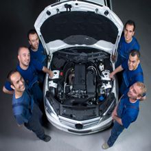 Six mechanics around a car with an open hood, viewed from above.