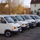 Fünf weiße Transporter parken nebeneinander auf einem Parkplatz vor einem Wohnhaus.
