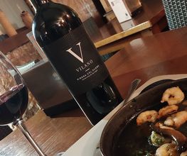 Botella de vino tinto y copa junto a un plato de camarones en una mesa de madera.