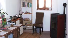 Ein kleiner Raum mit Tisch, braunem Stuhl, Regal, Büchern, einem Ofen und einem Fenster.