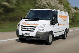 Weißer Lieferwagen fährt auf einer Landstraße, mit Bäumen im Hintergrund.