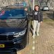 Een man staat naast een zwarte auto met een kenteken, op een zonnige dag.
