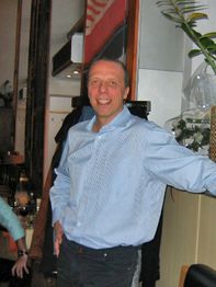 Ein lächelnder Mann in einem blauen Hemd steht in einem Raum neben einer Pflanze.