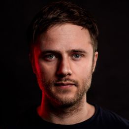 Porträt eines Mannes mit Bart, rot-schwarz beleuchtet, auf dunklem Hintergrund.