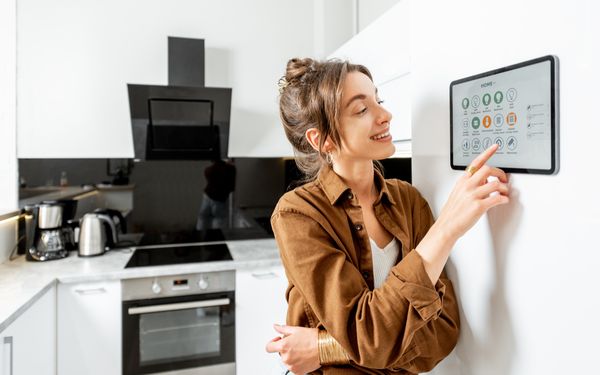 Eine Frau steht in ihrer Küche neben einem Tablet, welches an der Wand montiert wurde und bedient ihr Funk-Smart-Home System.