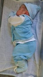 Bebê dormindo em um berço, vestido de azul, com gorro e meias combinando.