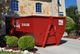 Red dumpster in a residential driveway beside a stone house with bushes in the foreground.