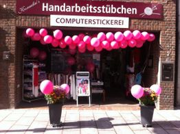 Außenansicht eines Handarbeitsstübchens mit rosa Luftballons, Blumen und Stickereischildern.