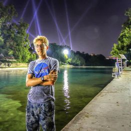Young man by a river at night, wearing a NASA shirt, with lights in the sky.