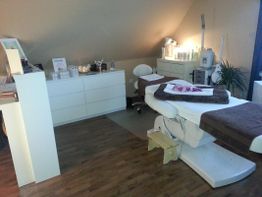 Kosmetikstudio mit Behandlungsstuhl, Handtücher und leuchtenden Lampen auf Holzboden.