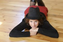 Frau mit Hut liegt lächelnd auf Holzboden, roter Rock und schwarzes Oberteil.