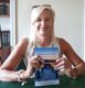 Una donna sorride, seduta a un tavolo, e tiene un libro intitolato "Ipnosi e Mindfulness".