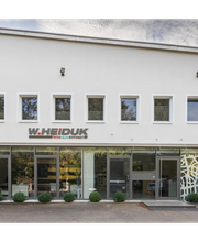Moderne weiße Gebäudefassade mit sechs Fenstern und einem Firmenschild, umgeben von Bäumen.