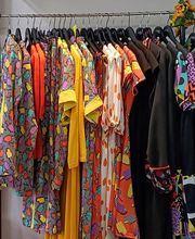 Ropa colorida colgada en un perchero con un estante encima que tiene un bolso y un jarrón.