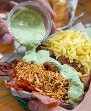 Manos vertiendo salsa verde sobre plato con pollo, tocino y queso rallado.